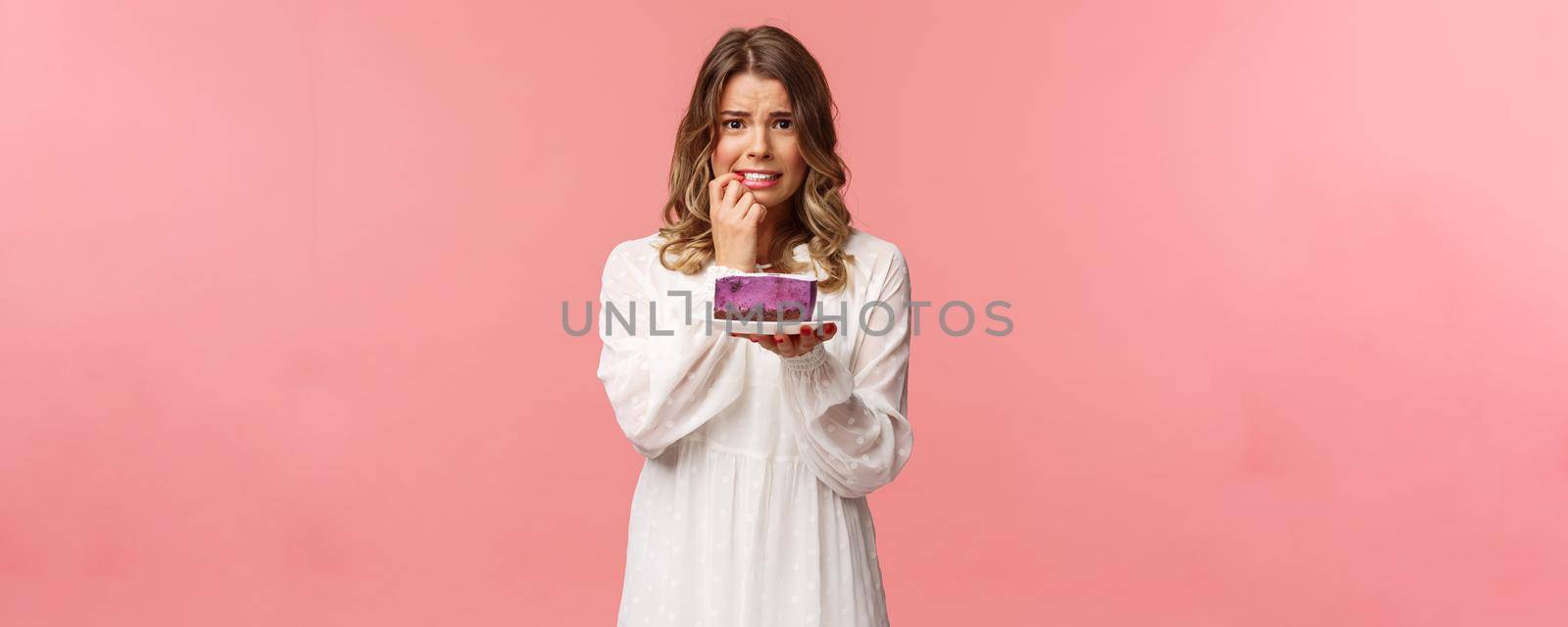 Holidays, spring and party concept. Alarmed, worried young blond girl hesitating eat cake or not, being on diet, trying stay healthy, holding dessert, biting finger nervously, pink background by Benzoix