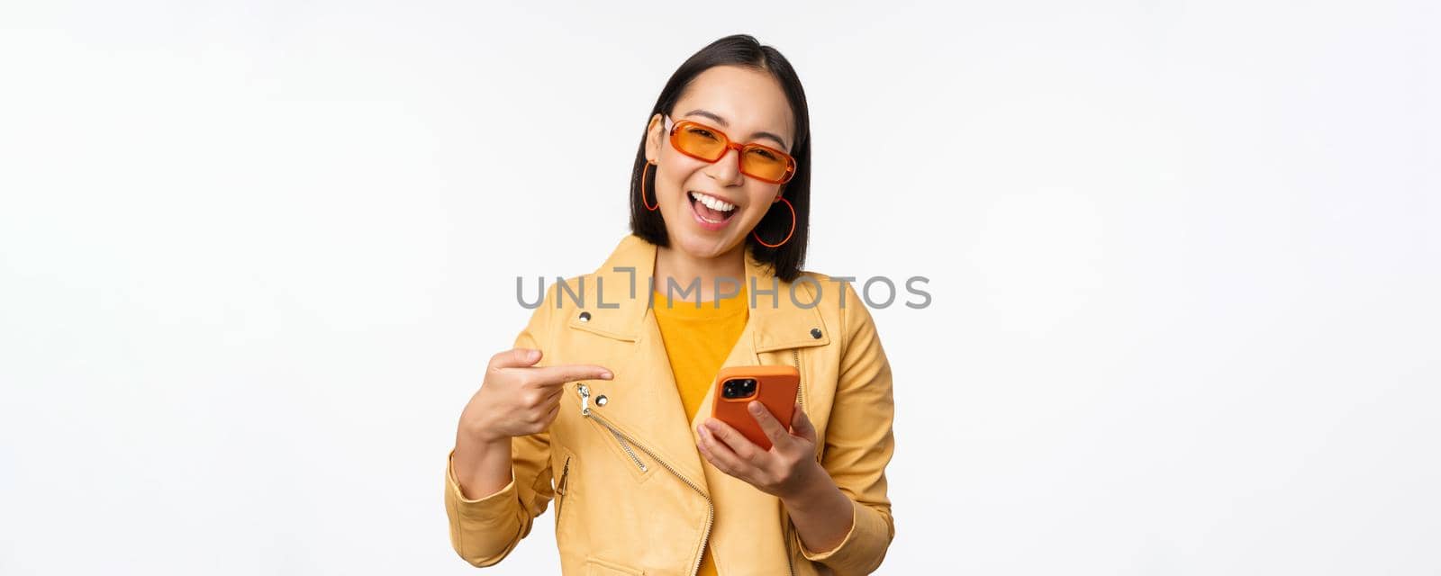 Happy smiling korean girl in sunglasses, holding smartphone, pointing at mobile phone screen, standing over white background by Benzoix