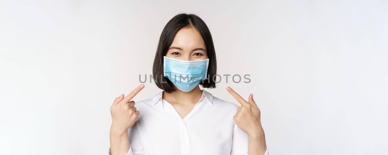 Image of young asian woman pointing at herself while wearing medical face mask, concept of covid-19 protection, standing over white background.