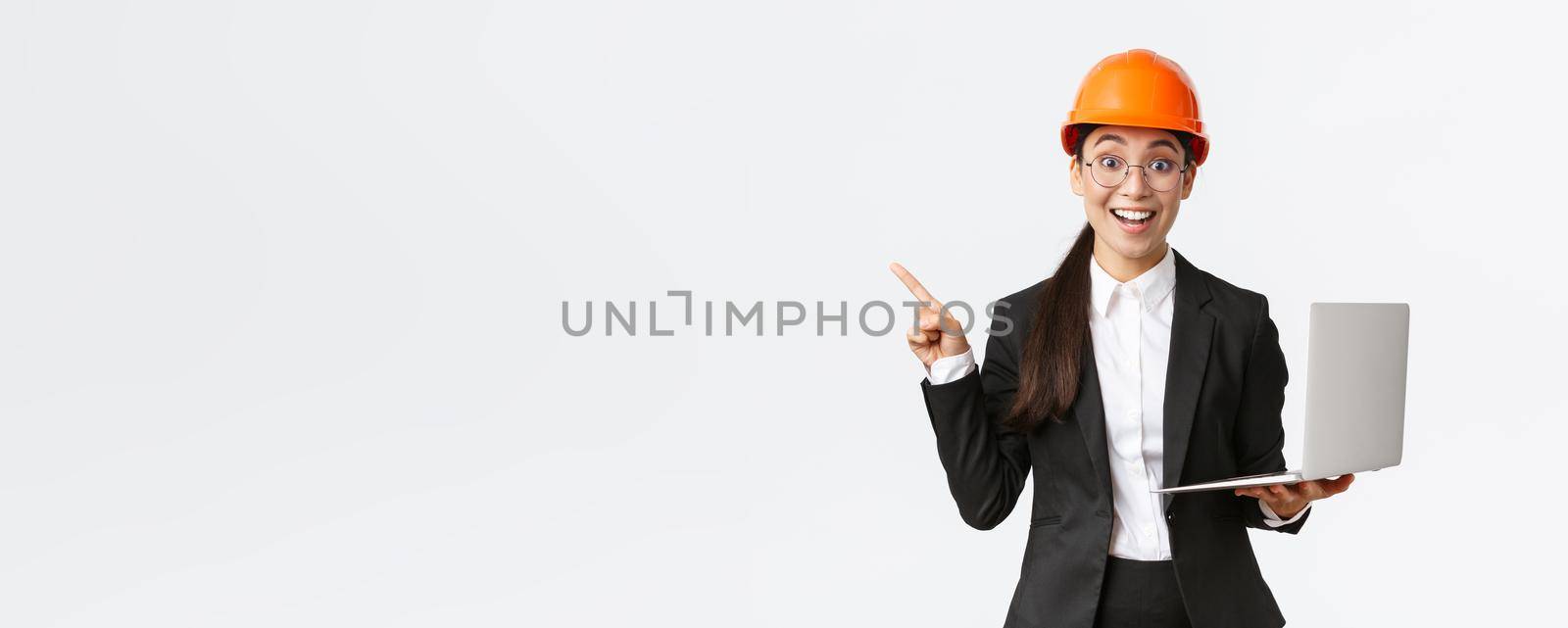 Excited happy asian female engineer, industrial woman in safety helmet and business suit, showing presentation, pointing finger at graph or chart and holding laptop computer, smiling amazed by Benzoix