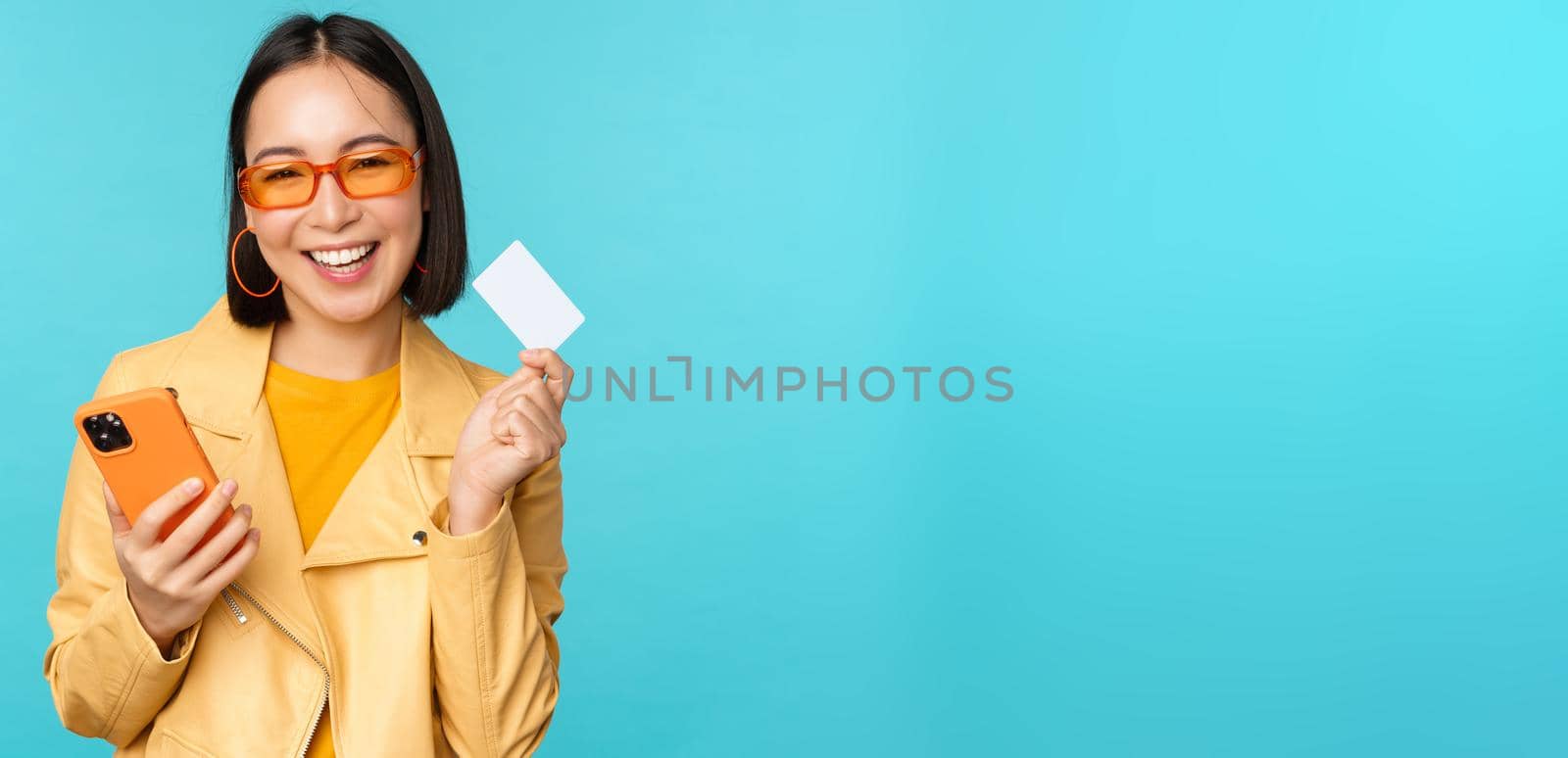 Online shopping. Stylish young asian woman in sunglasses, showing credit card and using smartphone, paying in internet, making purchase, standing over blue background.