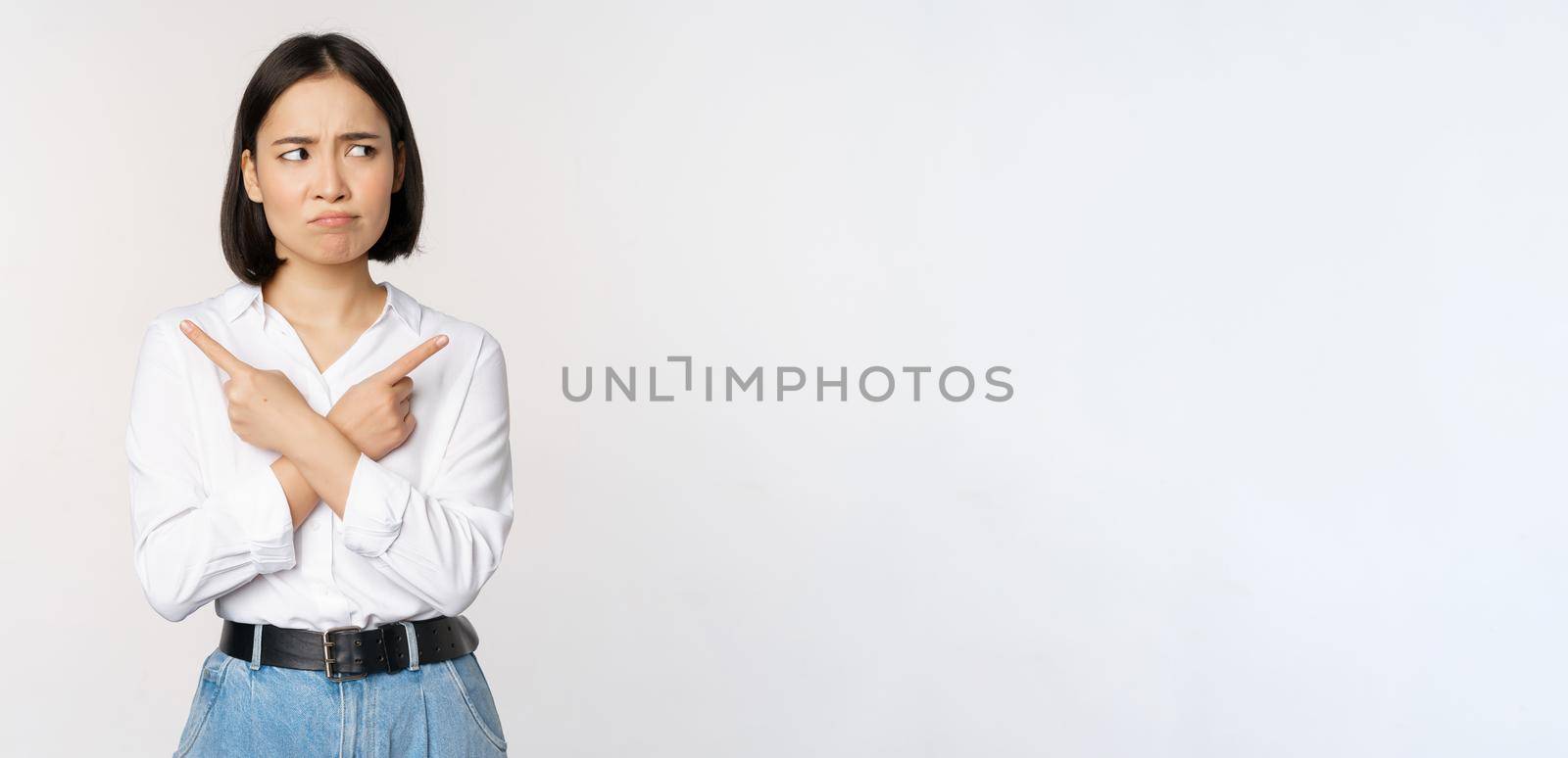 Indecisive asian woman pointing fingers sideways, pointing fingers and looking clueless, confused with choices, standing over white background.