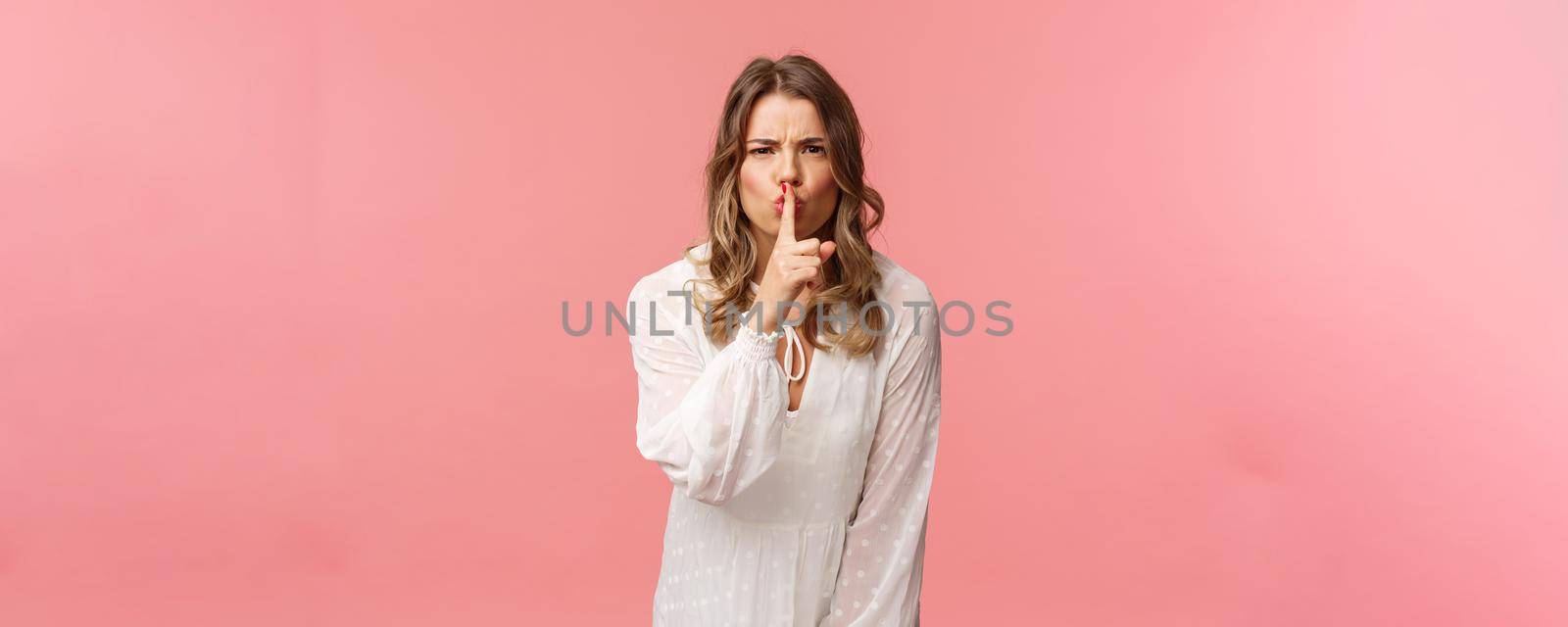 Beauty, fashion and women concept. Angry serious-looking blond girl in white dress, shushing at someone making noise, press finger to lips scolding bad behaviour, say be silent, pink background.