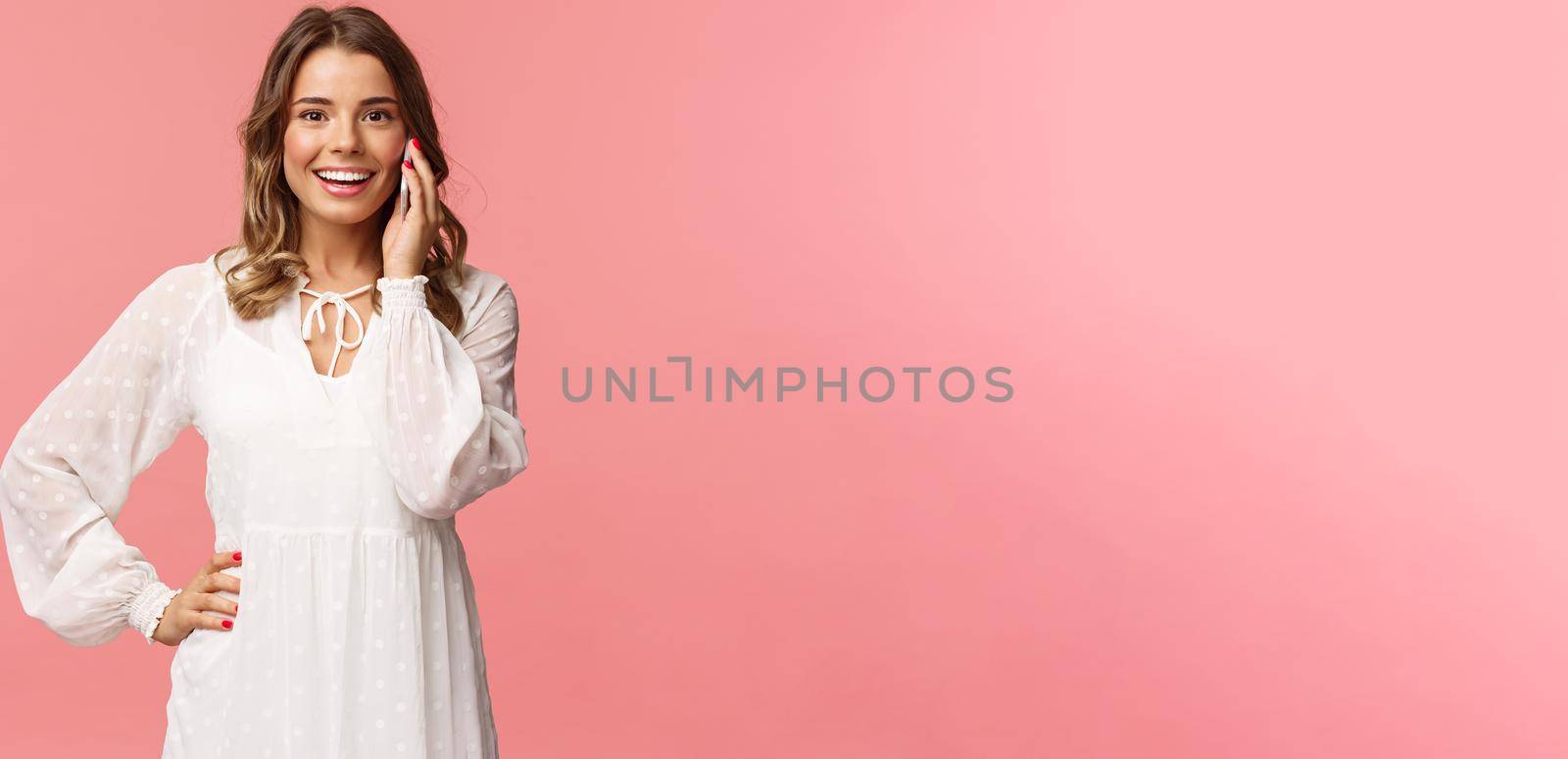 Portrait of joyful good-looking blond woman in white dress, talking on phone, hold smartphone near ear and look upbeat camera, smiling discuss friends date, stand pink background.