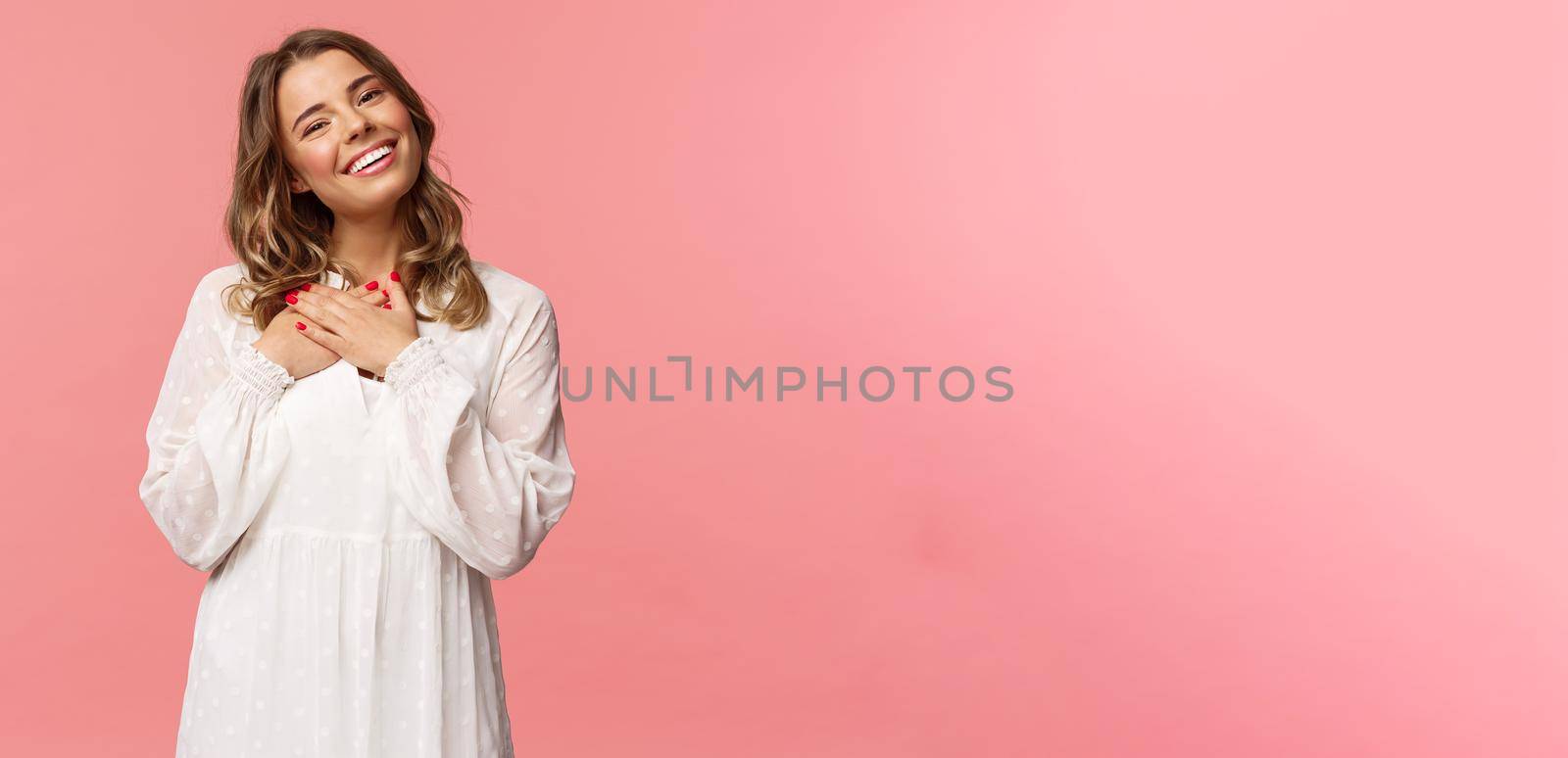 Beauty, fashion and women concept. Portrait of feminine blond young woman in white cute dress, hold arms pressed to heart and sighing from admiration and delight, feel thankful, pink background.