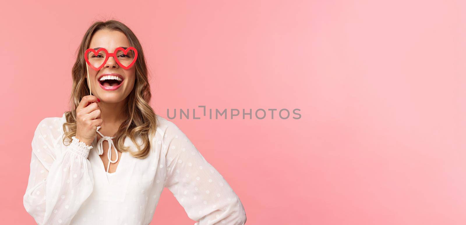 Spring, happiness and celebration concept. Close-up portrait of funny and carefree, beautiful caucasian woman with blond hair, white dress, holding heart-shaped glasses mask and laughing.