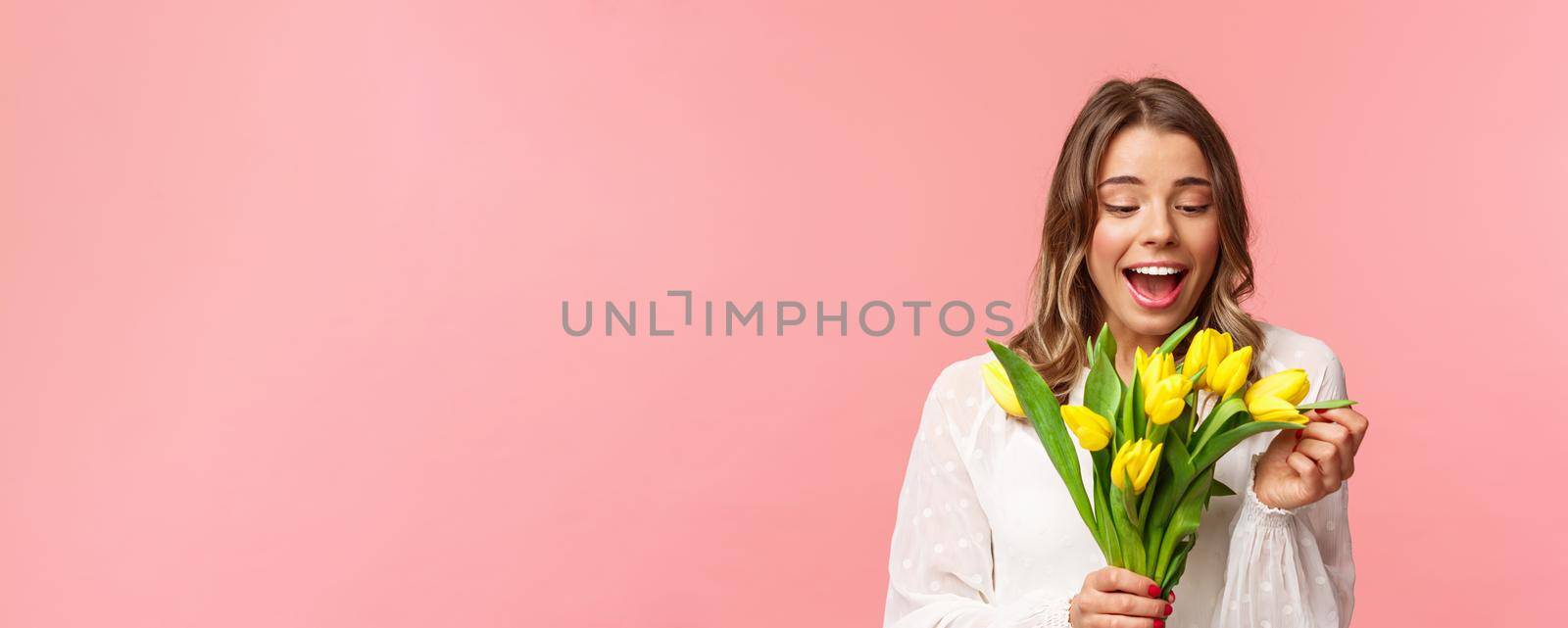 Spring, happiness and celebration concept. Portrait of surprised and pleased happy girl cheerfully looking at bouquet of flowers, smiling amused, receive yellow tulips, stand pink background by Benzoix