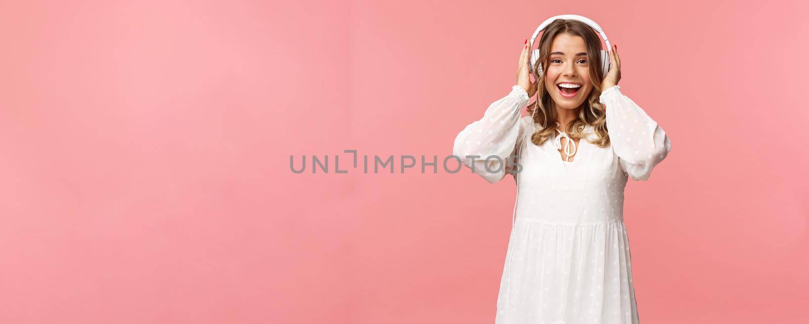 Portrait of excited, happy good-looking girl in white tender dress, wearing headphones and smiling amazed as looking at camera, fascinated with good sound quality, pink background by Benzoix