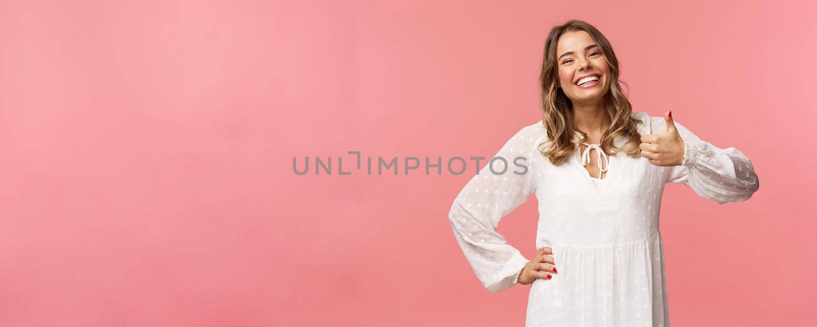 Enthusiastic lovely young woman in white cute dress, show thumb-up in approval, recommend product, smiling satisfied, leave positive review, impression, standing pink background.