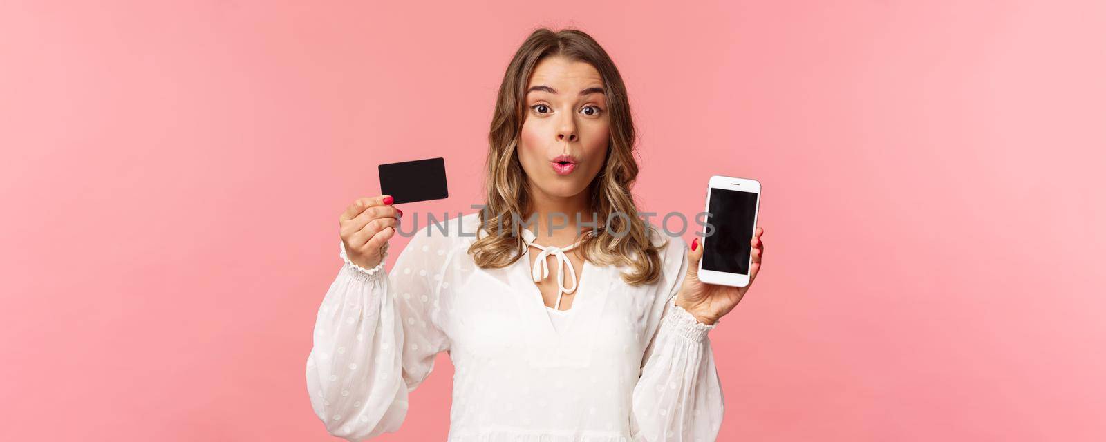 Finance, shopping and technology concept. Close-up portrait of excited blond cute girl in white dress, fold lips amused look camera as showing credit card and mobile phone, pink background by Benzoix