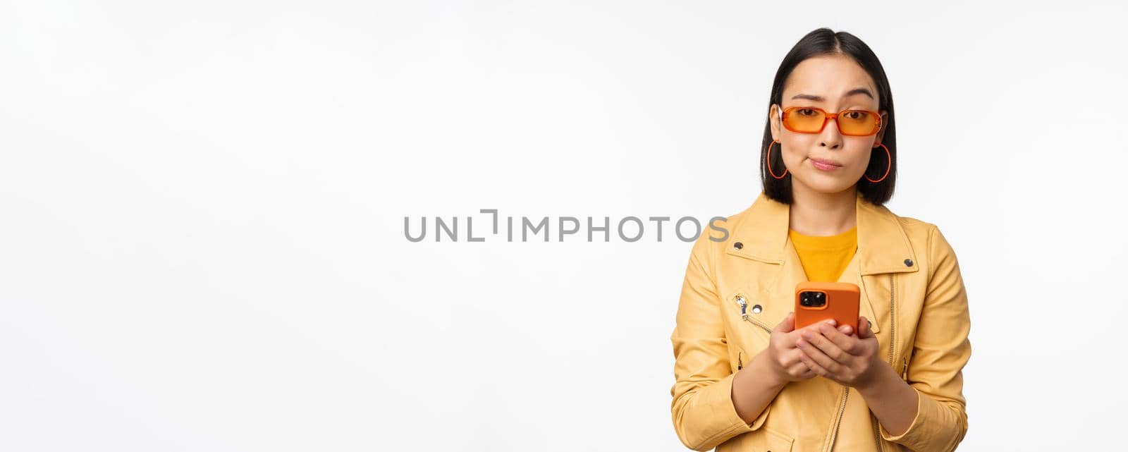 Portrait of confused asian girl in sunglasses, holding smartphone, using mobile phone and looking puzzled, standing clueless over white background by Benzoix