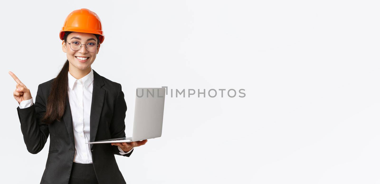 Smiling professional asian female engineer or architect at construction, wearing safety helmet and suit, pointing finger left while using laptop computer, standing white background.