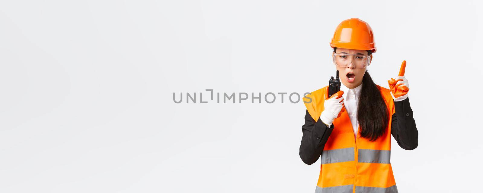 Serious-looking asian female industrial engineer, technician in safety helmet and uniform command construction team with walkie-talkie, explain or scolding someone with radio phone by Benzoix
