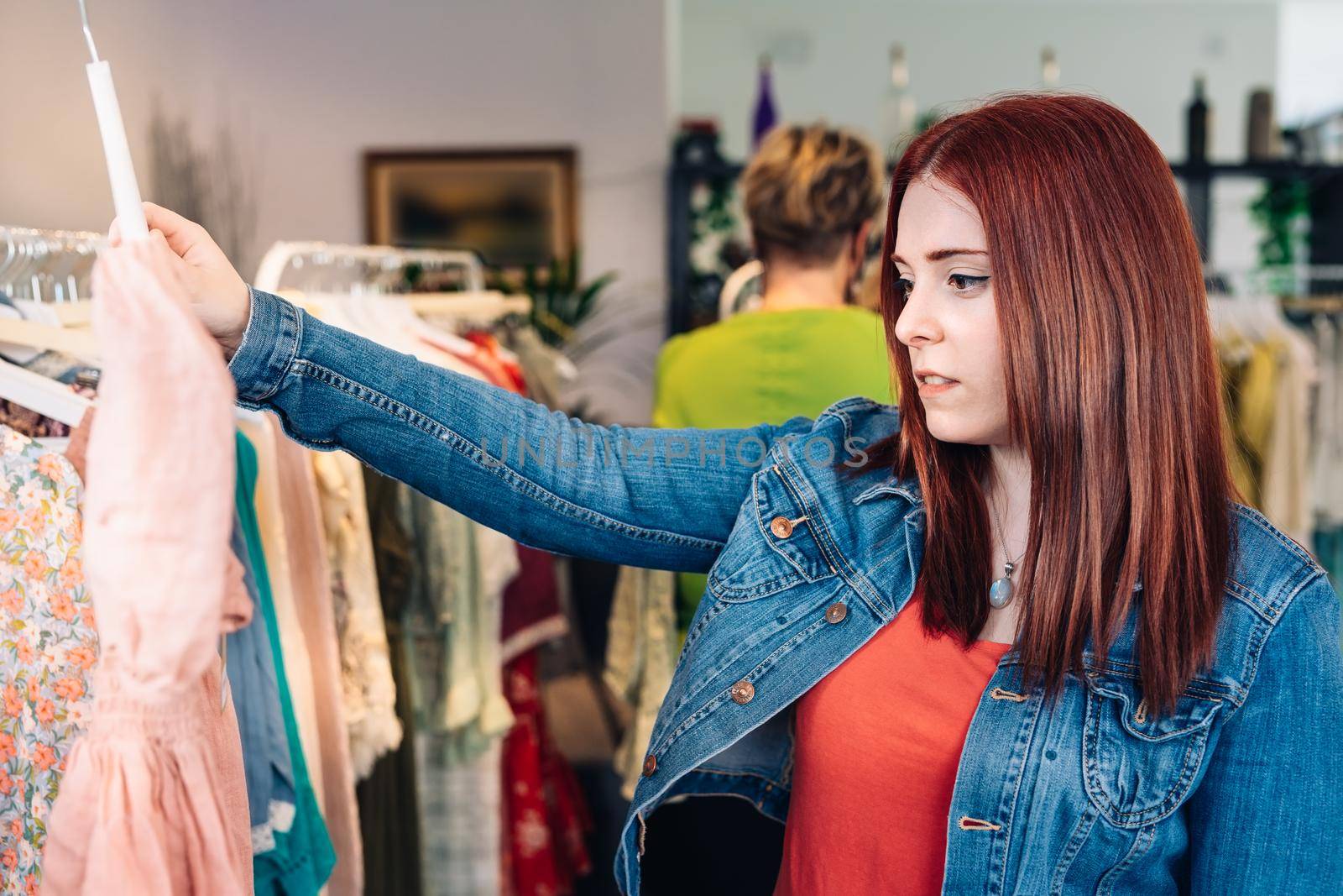 young red-haired woman choosing clothes from a rack, shopping in a fashion shop. shopping concept. by CatPhotography