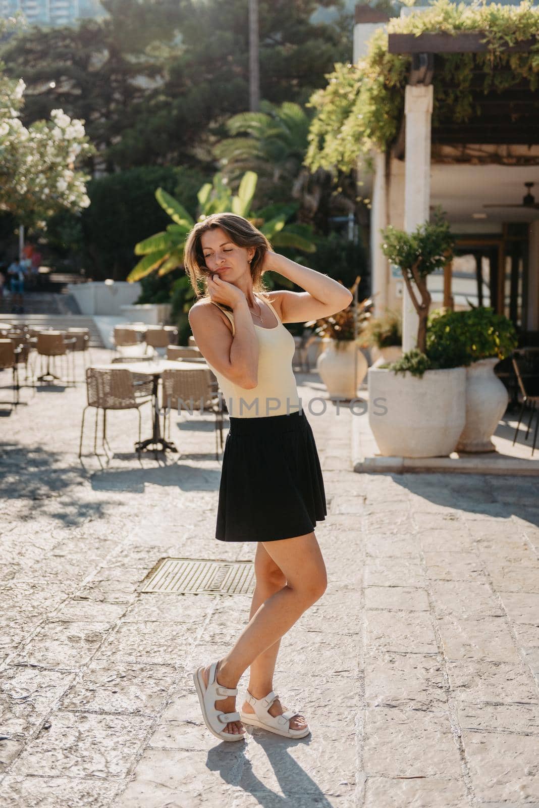 Girl Tourist Walking Through Ancient Narrow Street On A Beautiful Summer Day In MEDITERRANEAN MEDIEVAL CITY, OLD TOWN BUDVA, MONTENEGRO. Young Beautiful Cheerful Woman Walking On Old Street At Tropical Town. Pretty Girl Looking At You And Smiling by Andrii_Ko