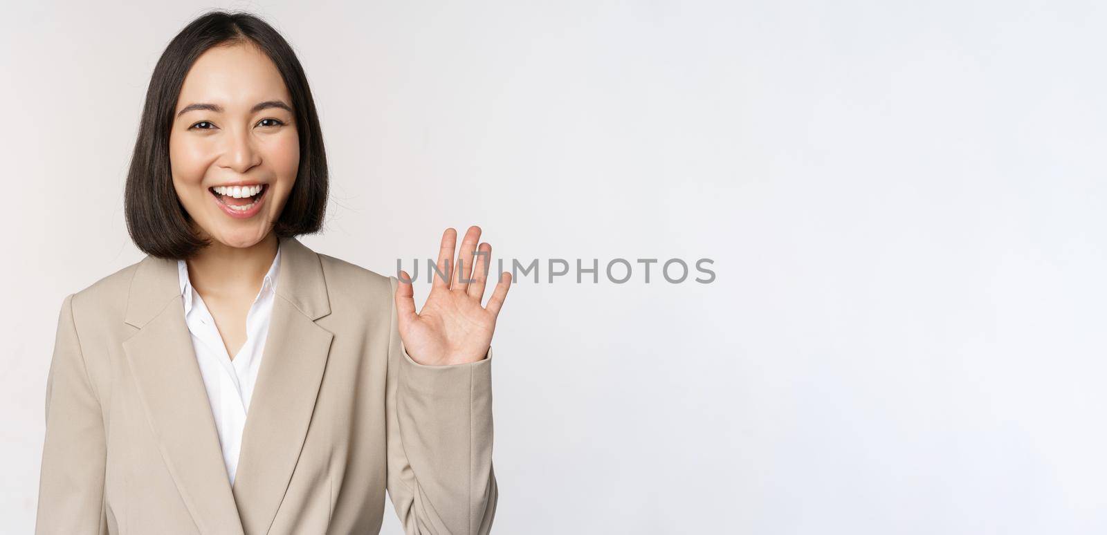 Friendly business woman, asian office lady waving hand and saying hello, hi gesture, standing over white background by Benzoix