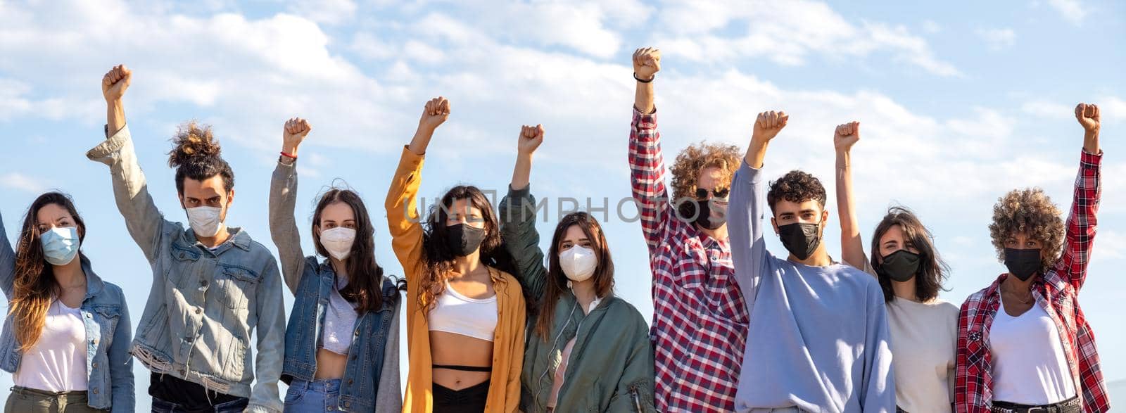 Panoramic image of multiracial activist protesters with fists raised up in the air wearing protective face mask protesting on the street. Demonstration concept.