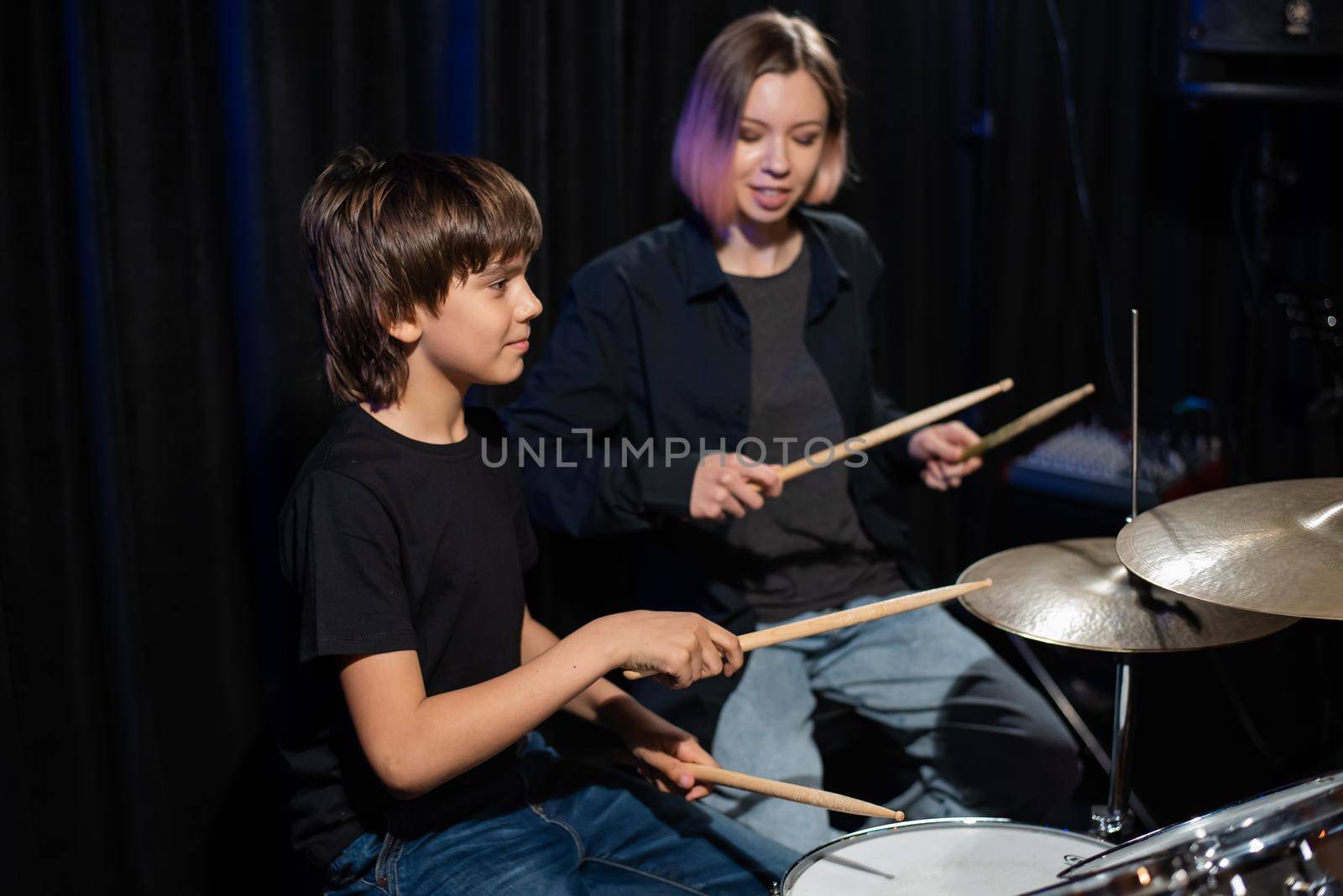 Young caucasian woman teaches a boy to play the drums in the studio on a black background. Music school student by mrwed54