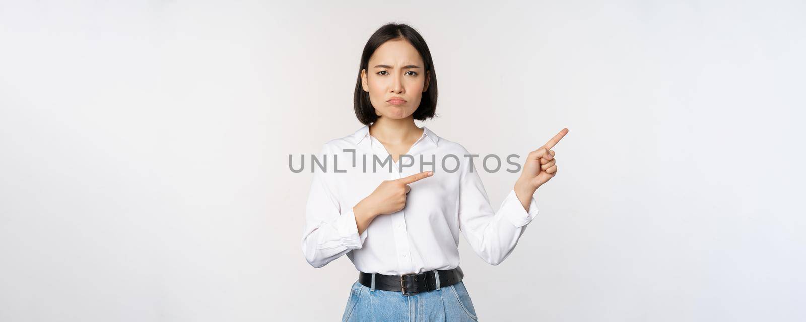 Sad and gloomy asian woman looking disappointed, complaining at banner or advertisement, pointing fingers right at promo and frowning upset, white background by Benzoix