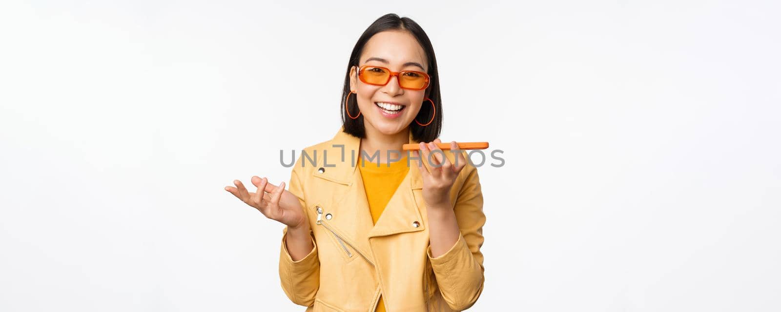 Image of happy asian girl talking on speakerphone, recording, translating her voice with mobile phone app, talking in smartphone dynamic, standing over white background.