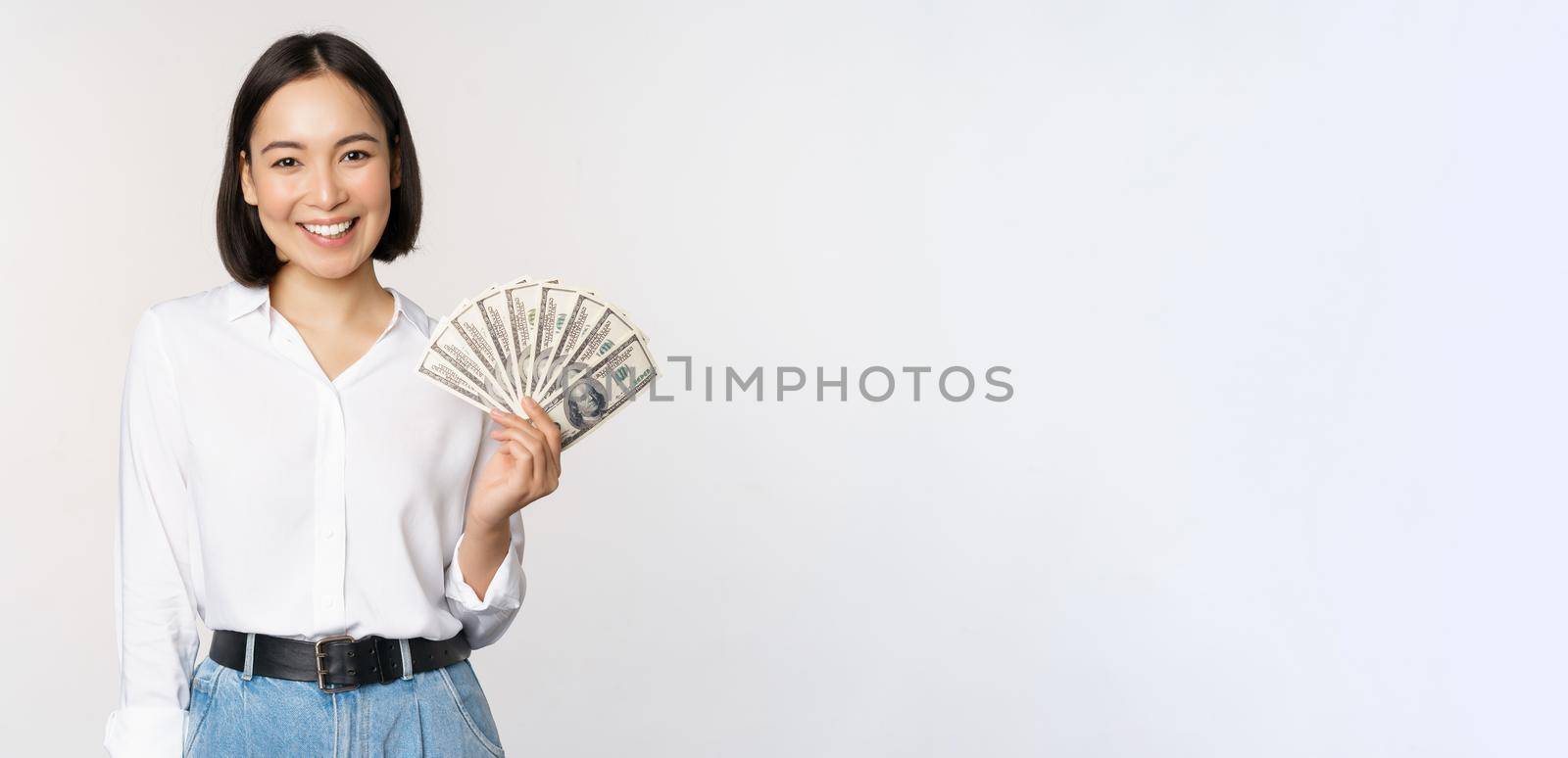 Credit and loan concept. Smiling young asian woman holding cash dollars and looking happy at camera, white background.