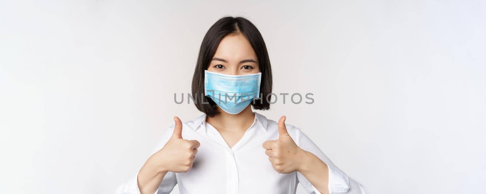 Portrait of smiling asian office lady in medical face mask, showing thumbs up, recommending smth, standing over white background by Benzoix