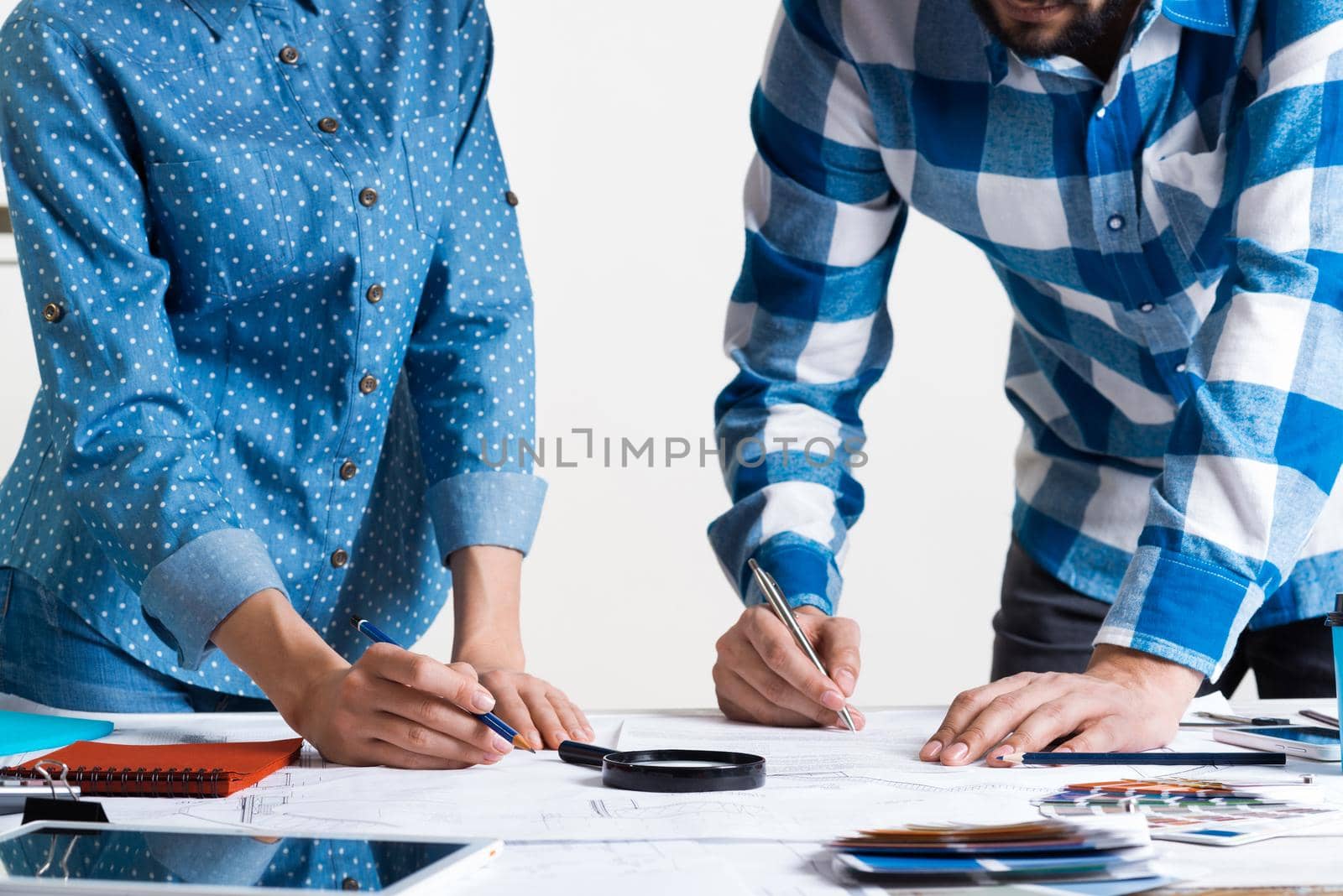 Man carefully studying technical drawing with magnifying glass. Designers working with color swatches and construction blueprint at workplace. Thorough analysis and inspection of design project.