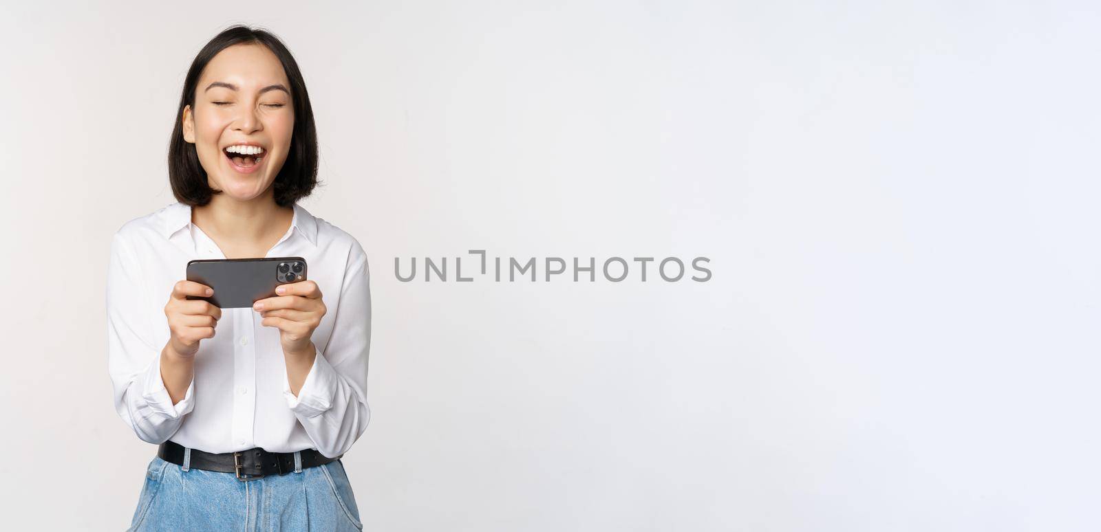 Image of young asian woman watching on smartphone app, holding mobile phone and looking at screen, laughing and smiling, standing over white background by Benzoix