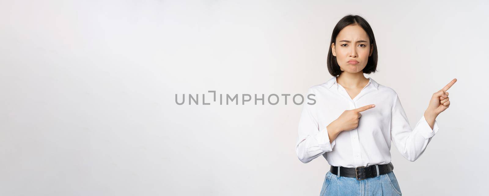 Sad and gloomy asian woman looking disappointed, complaining at banner or advertisement, pointing fingers right at promo and frowning upset, white background by Benzoix