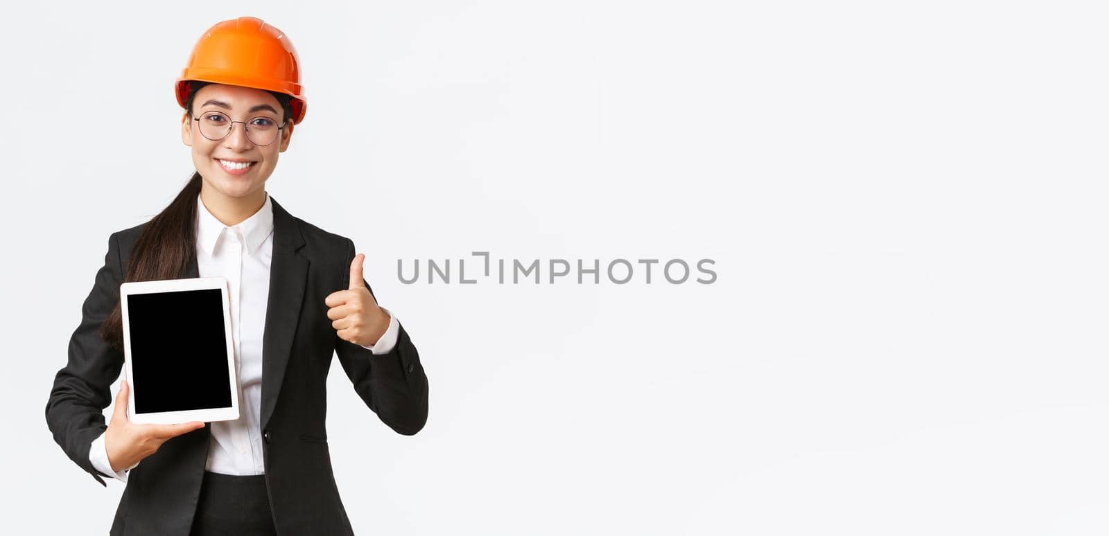 Professional smiling asian female construction manager, factory engineer in business suit and safety helmet showing thumbs-up and digital tablet screen with positive graph, white background.