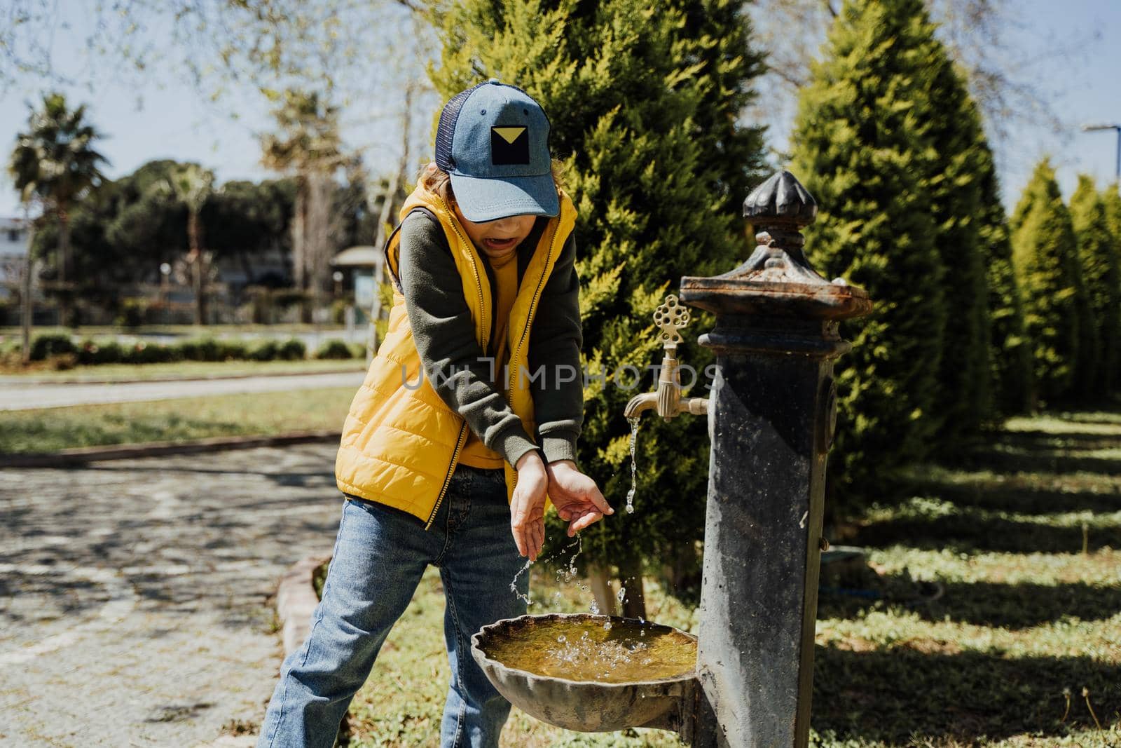 Kid schoolboy in casual clothing washing his hands in the street old fashioned drinking water fountain. Boy in blue jeans and yellow vest playing with water from drinking sprinkler in city park.