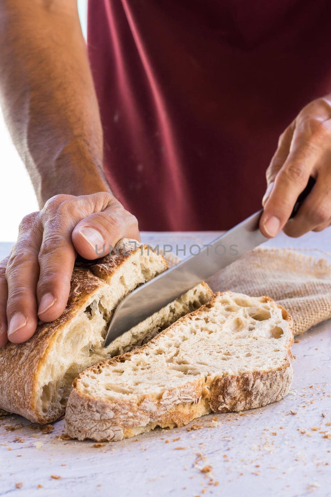 vertical image of man's hands breaking organic bread in the foreground with knife surrounded by crumbs on white table with natural light