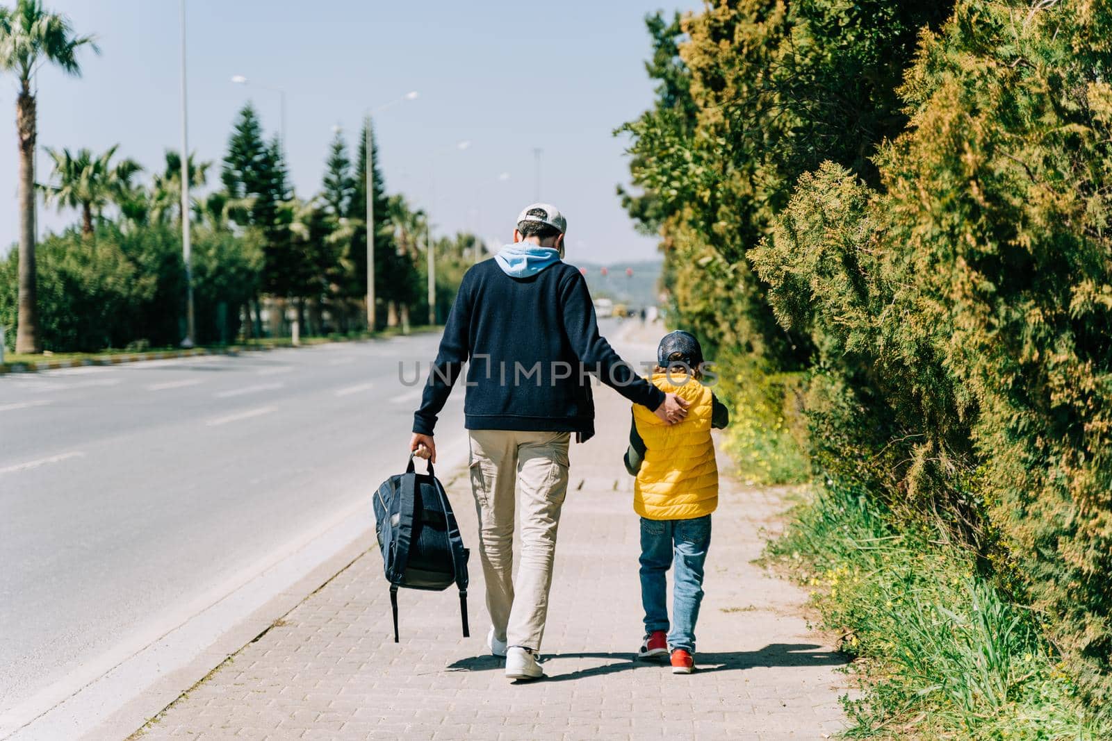 Rearview of father and son walking near city road. Dad holding his kid's back when taking a stroll in the southern city. Man and boy going to school near highway.