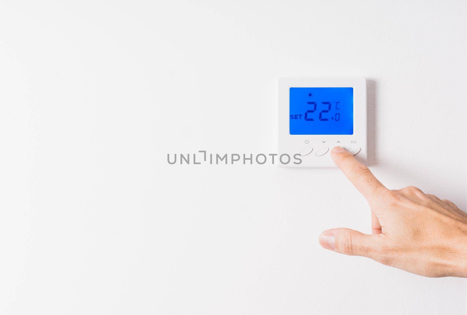 Caucasian man's hand adjusting the temperature of a white thermostat to twenty-two degrees with the blue backlit display on a white wall