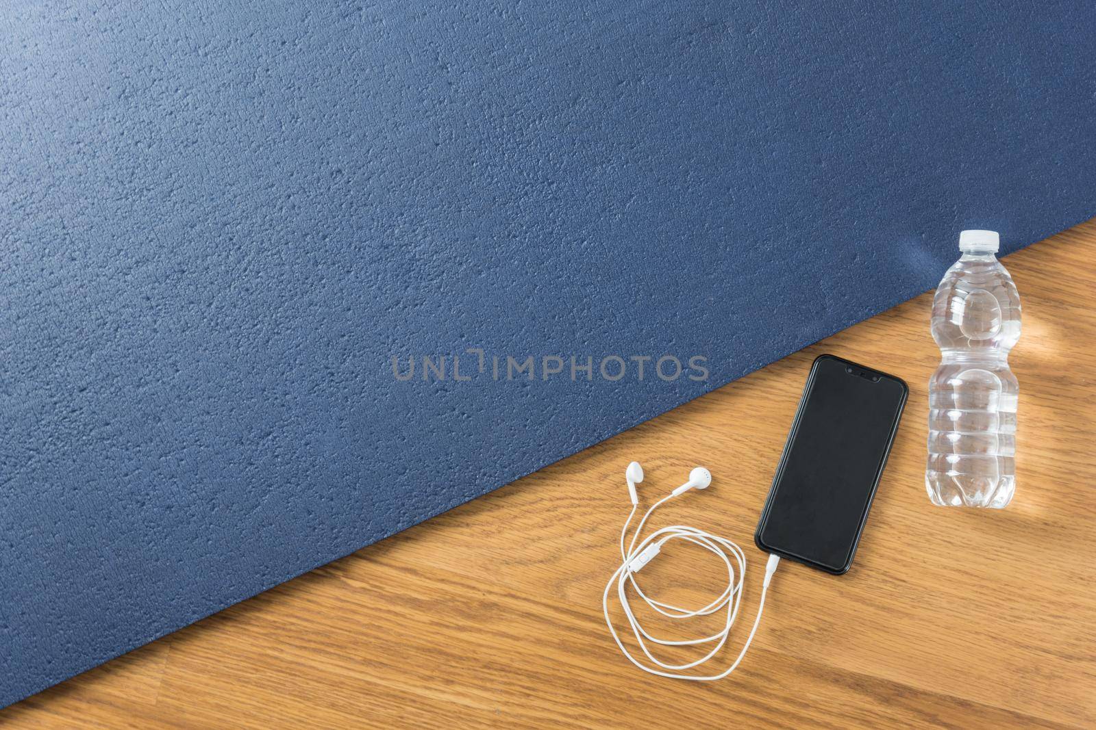 Image from above blue exercise mat at home with smartphone water bottle and white headphones on brown wooden floor
