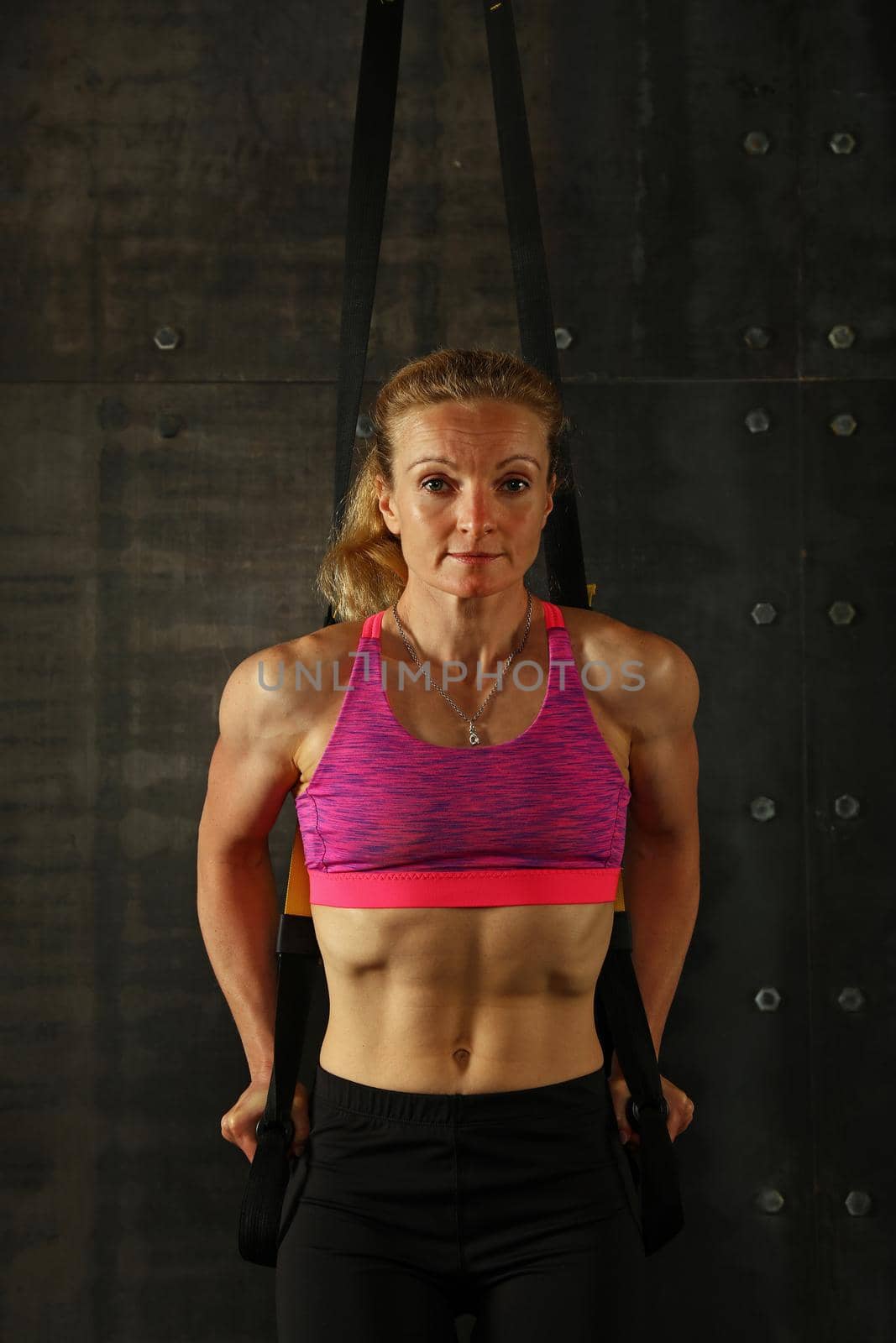 One young middle age athletic woman at crossfit training, exercising with trx suspension fitness straps over dark background, front view, looking at camera