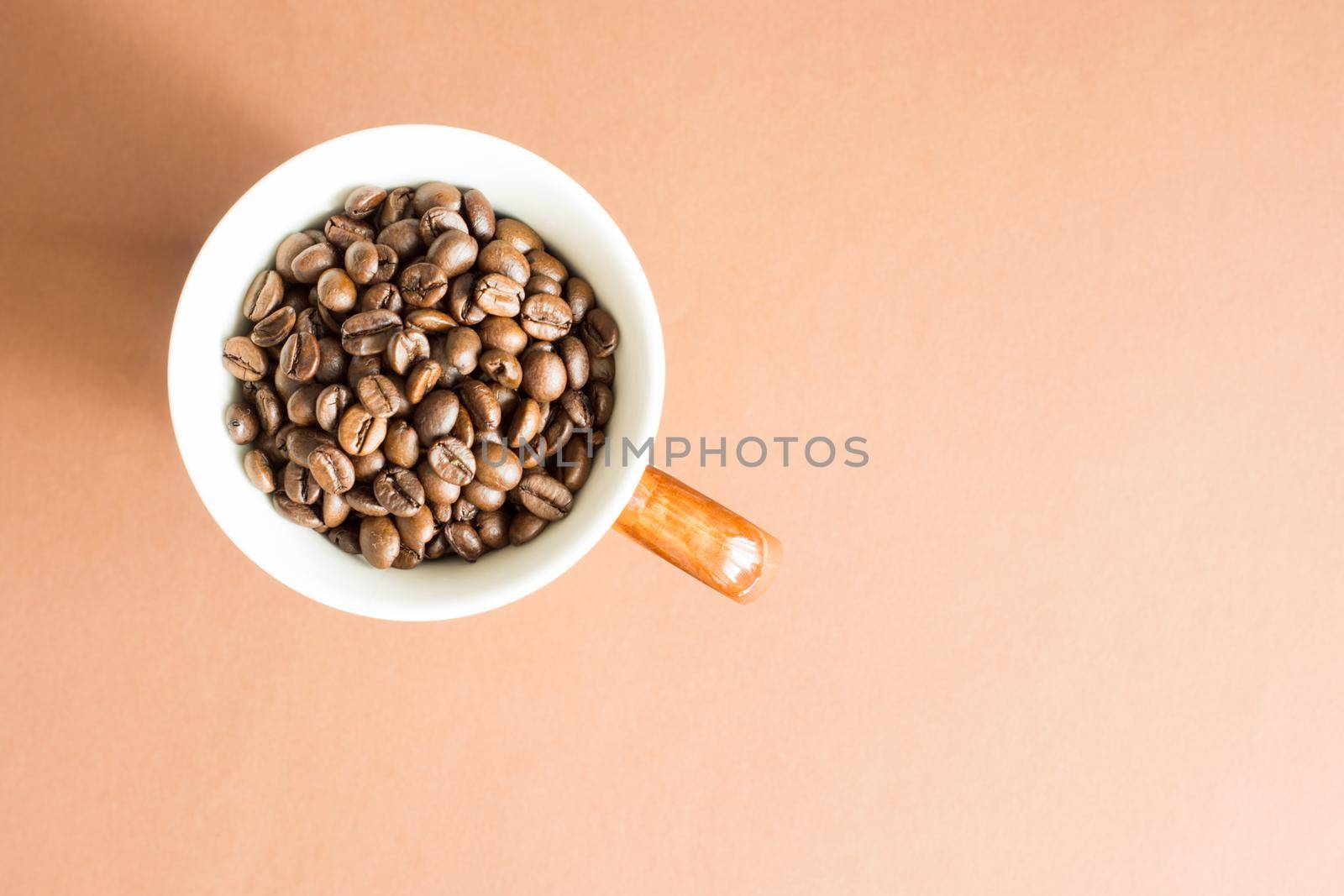 White cup with brown handle filled with coffee beans picked from above with light brown background