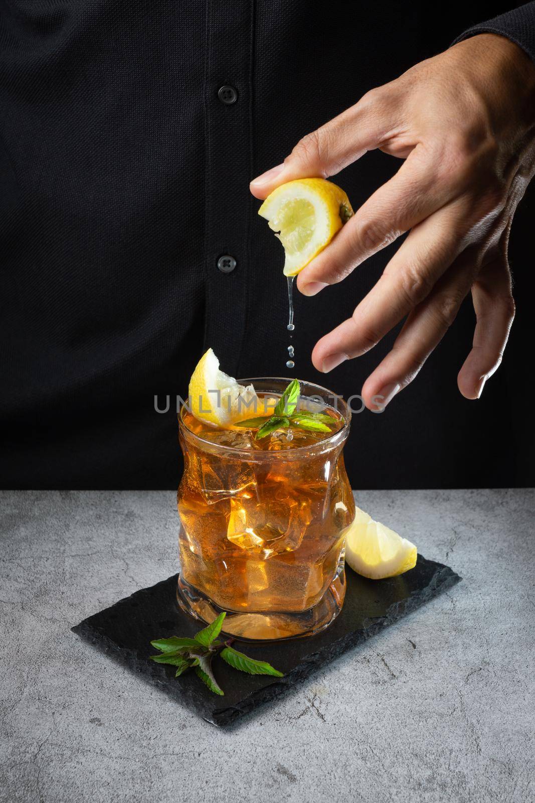 glass of tea with ice and lemon slices with fresh mint leaves and hand of the bartender in a black shirt squeezing a piece of lemon on top of the glass