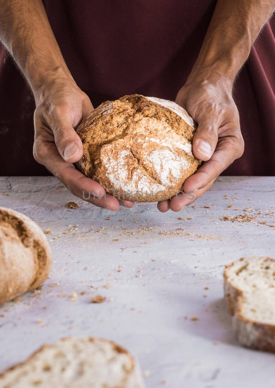 vertical image of man's hands holding an organic bread and in the foreground unfocused slices of bread surrounded by crumbs on white table with natural light