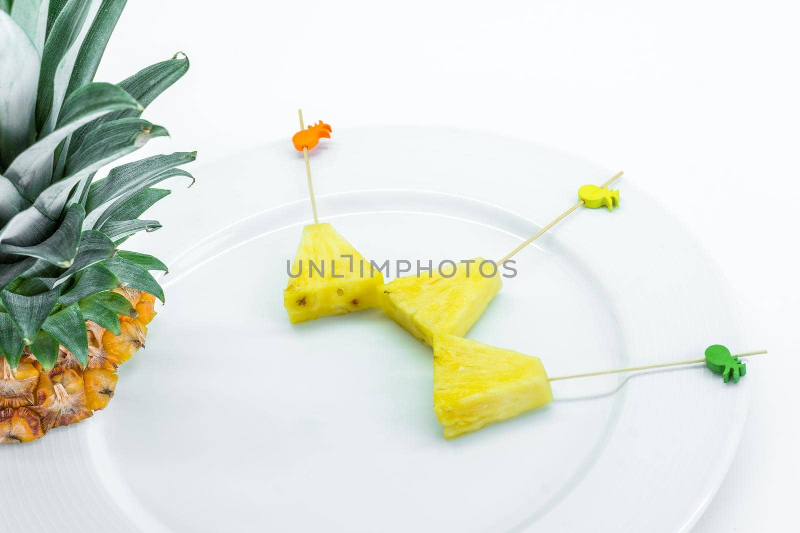 Pineapple skewers peeled and cut into triangles with chopsticks decorated on white plate with pineapple leaves on white background