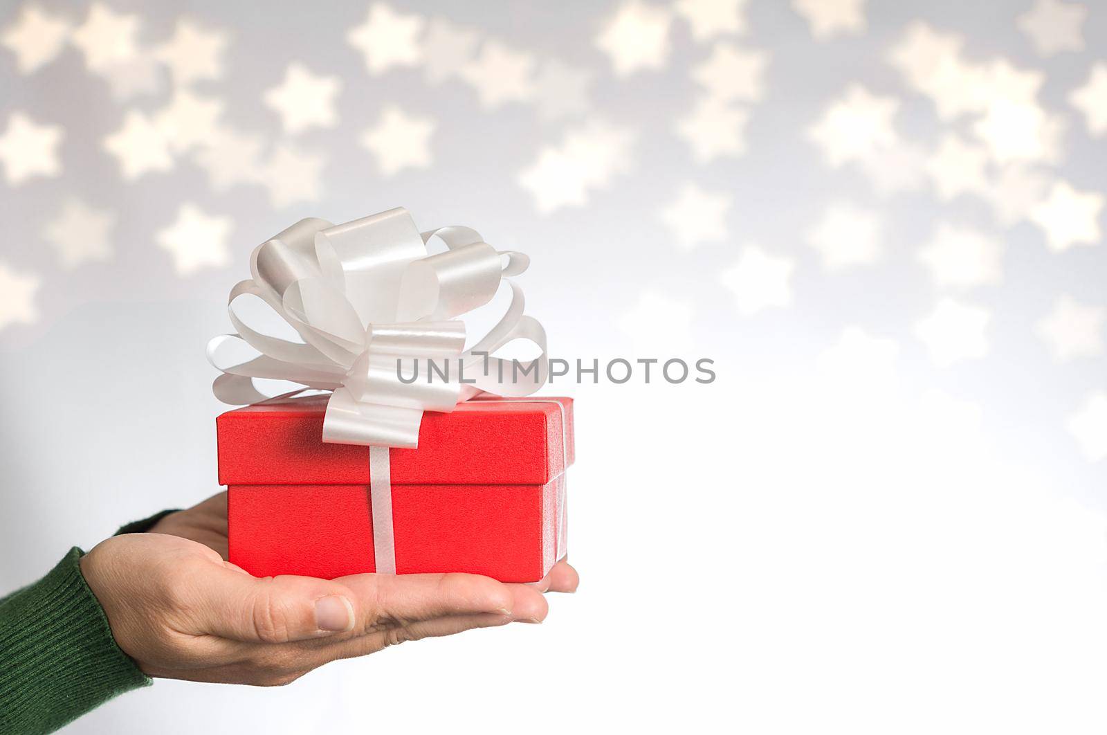 Hands of a woman in green sweater with red gift and white ribbon on neutral background with bokeh of stars