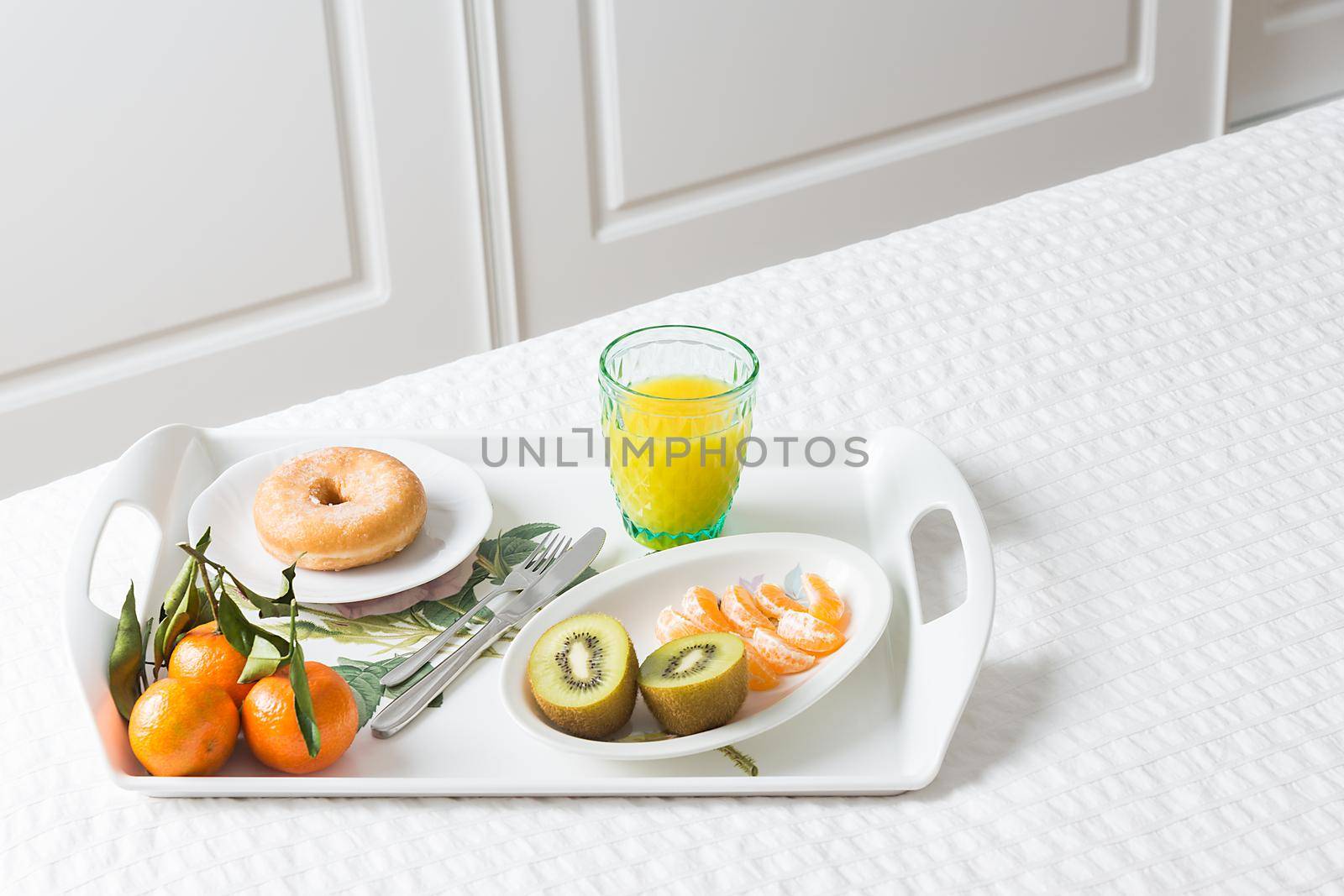 Horizontal image of a tray with a breakfast donut with mandarin kiwi and orange juice on a white bed with white background doors