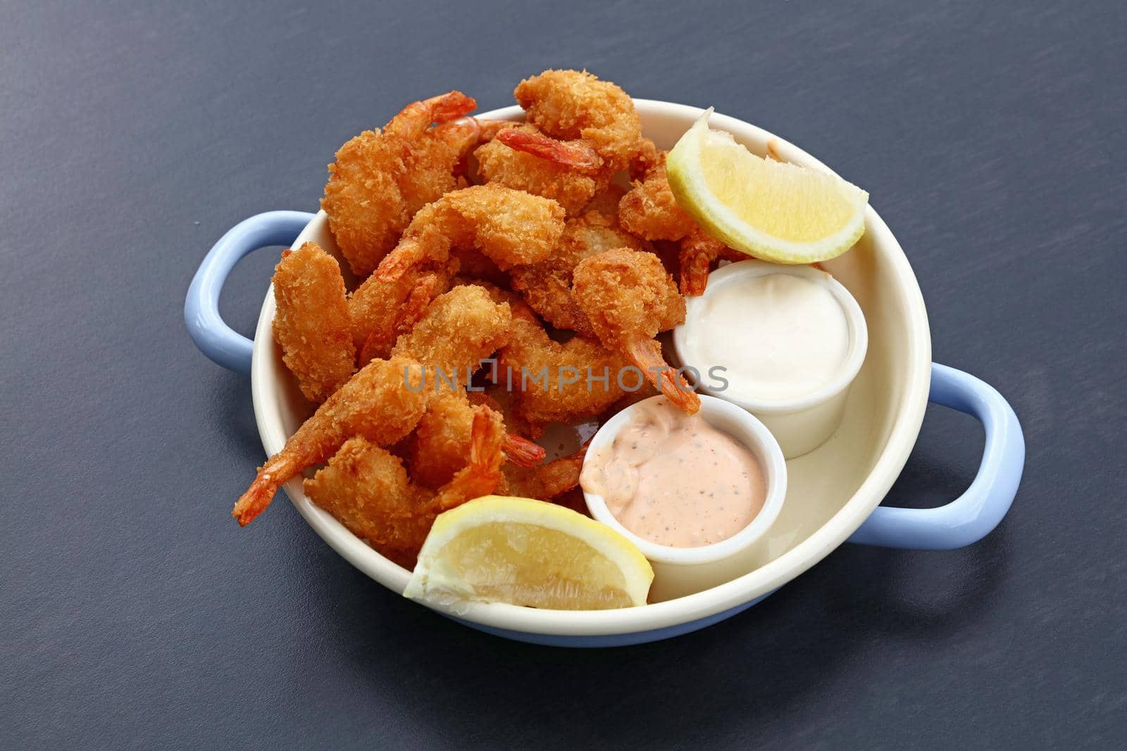 Close up portion of battered and breaded shrimps with dipping sauces served in cooking pot over blue table copy space, high angle view