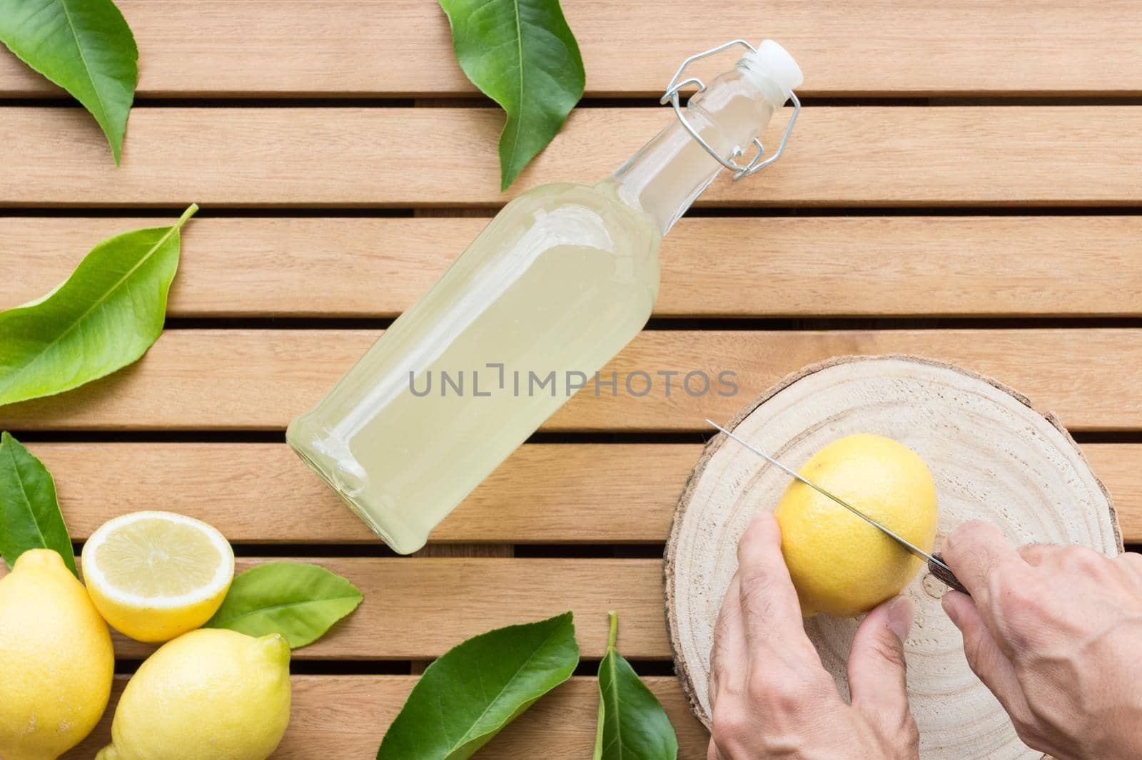 horizontal image of glass bottle full of lemonade with lemons in the lower left corner and hands cutting a lemon with a knife surrounded by lemon leaves