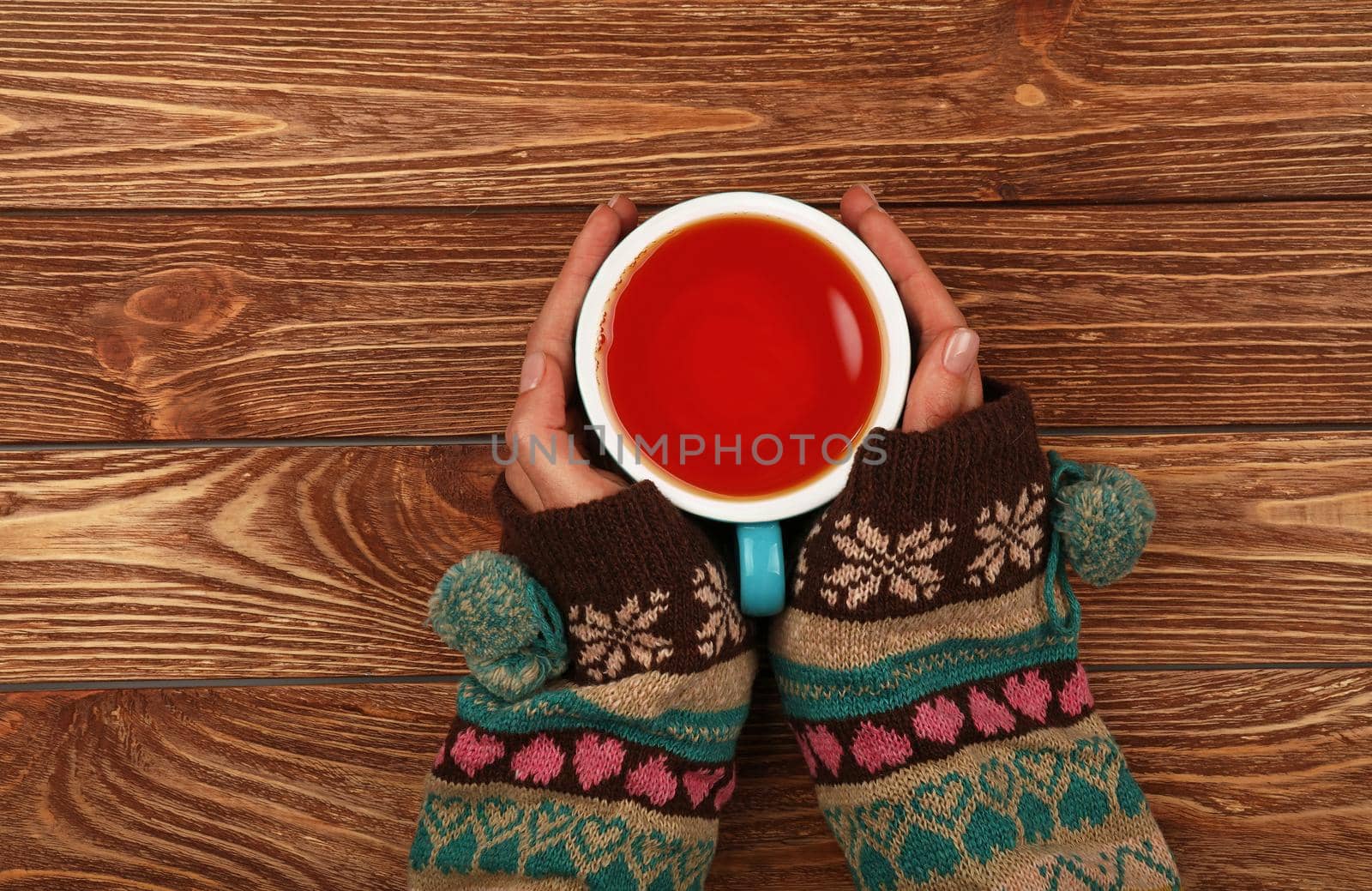 Close up two woman hands hold and hug big full cup of black tea or red fruit tea over brown wooden table, elevated top view, directly above