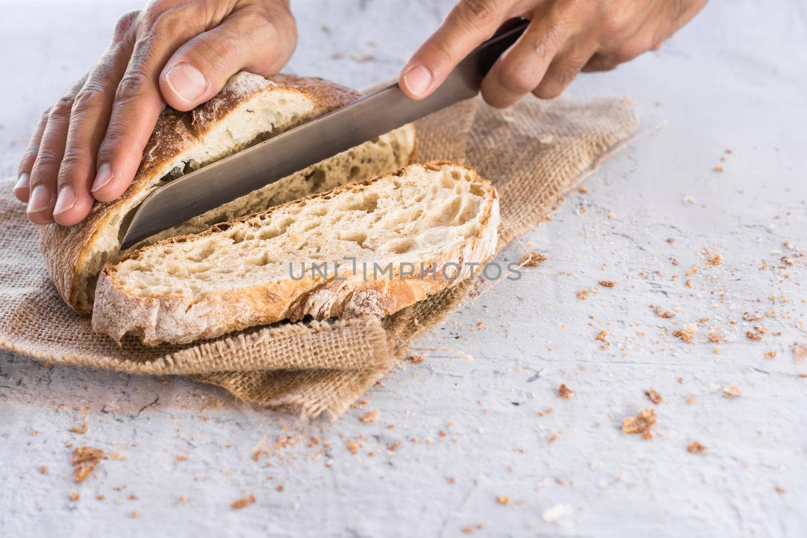 horizontal image of man's hands breaking organic bread in the foreground with knife surrounded by crumbs on white table with natural light