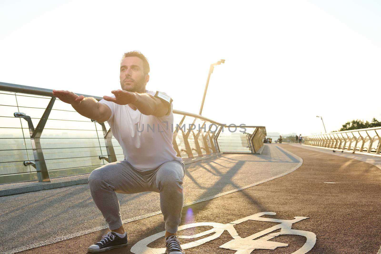 Middle aged Caucasian athlete doing deep squats on a city bridge on a sunny summer day. Concept of sport, movement, energy and dynamic, healthy lifestyle.