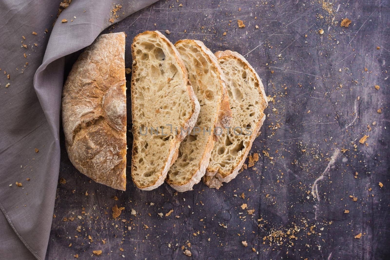 Zenithal image of an ecological rustic bread divided into half slices on a dark surface surrounded by crumbs and a gray cloth with natural light and a touch of flash