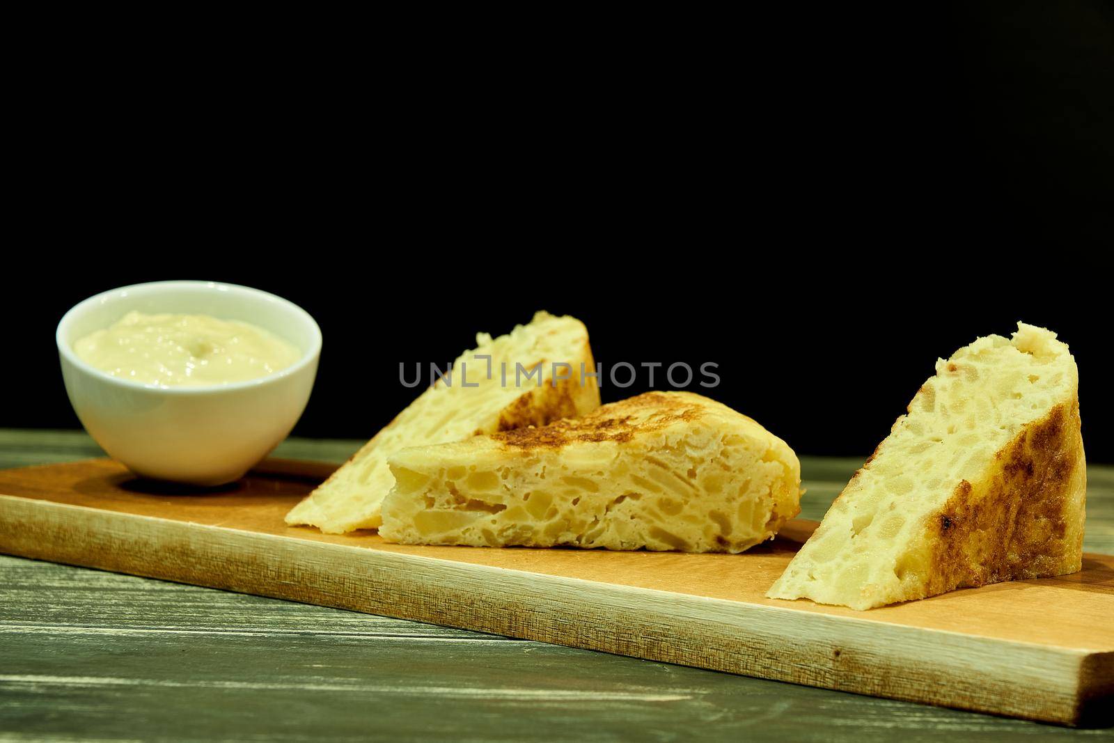 three skewers of potato omelette on an elongated brown wooden table with a bowl of white mayonnaise on a rustic wooden table and black background