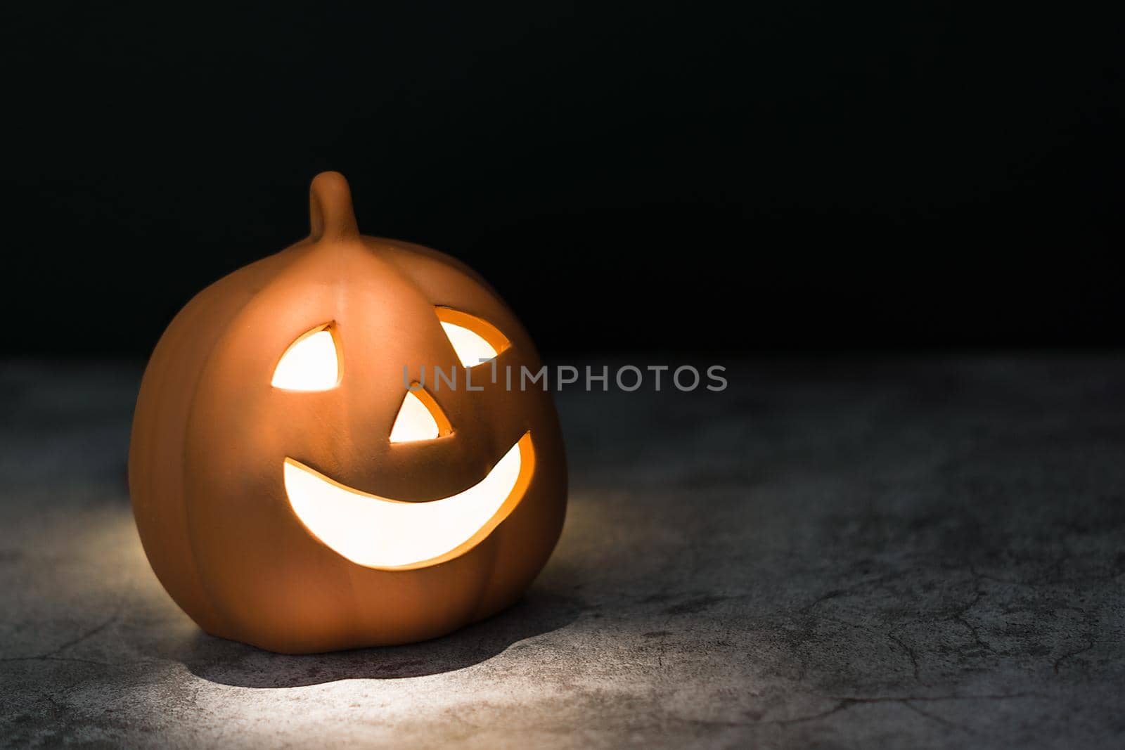Smiling ceramic Halloween pumpkin lit inside and from above with warm light on dark gray textured worn surface with black background and room for edit